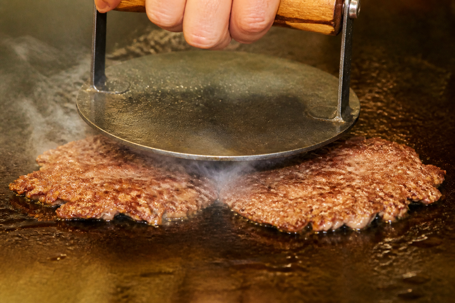 高温の鉄板で牛ひき肉を金属製プレスで薄く押し広げる様子